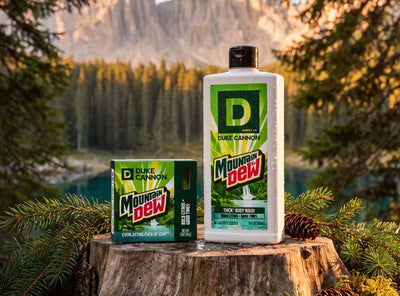 A bottle of Duke Cannon Mountain Dew Thick Body Wash and a matching box of Mountain Dew body soap are displayed on a tree stump with a forest and mountain background.