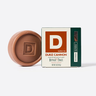 Puck shaped Bar of soap with Duke Cannon packaging on a white Background. Buffalo Trace inspired Bourbon scent. Bourbon - Oak Barrels + Aged Spirits scent.