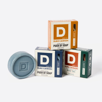 Duke Cannon soap pucks and packaging on a white background. Products shown: Midnight Swim Puck of Soap, Sawtooth Puck of Soap and Bourbon Puck of Soap.
