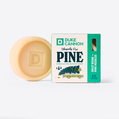 Duke Cannon Illegally Cut Pine Everlasting Puck of Soap next to it's packaging. On a white background. Scent is Splitwood + Pine Needles. 