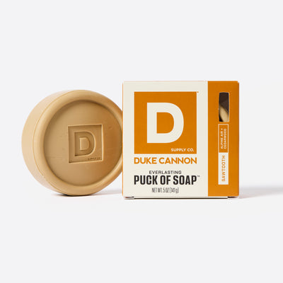Puck shaped Bar of soap with Duke Cannon packaging on a white Background. Sawtooth and Alpine Air + Cedarwood scent.